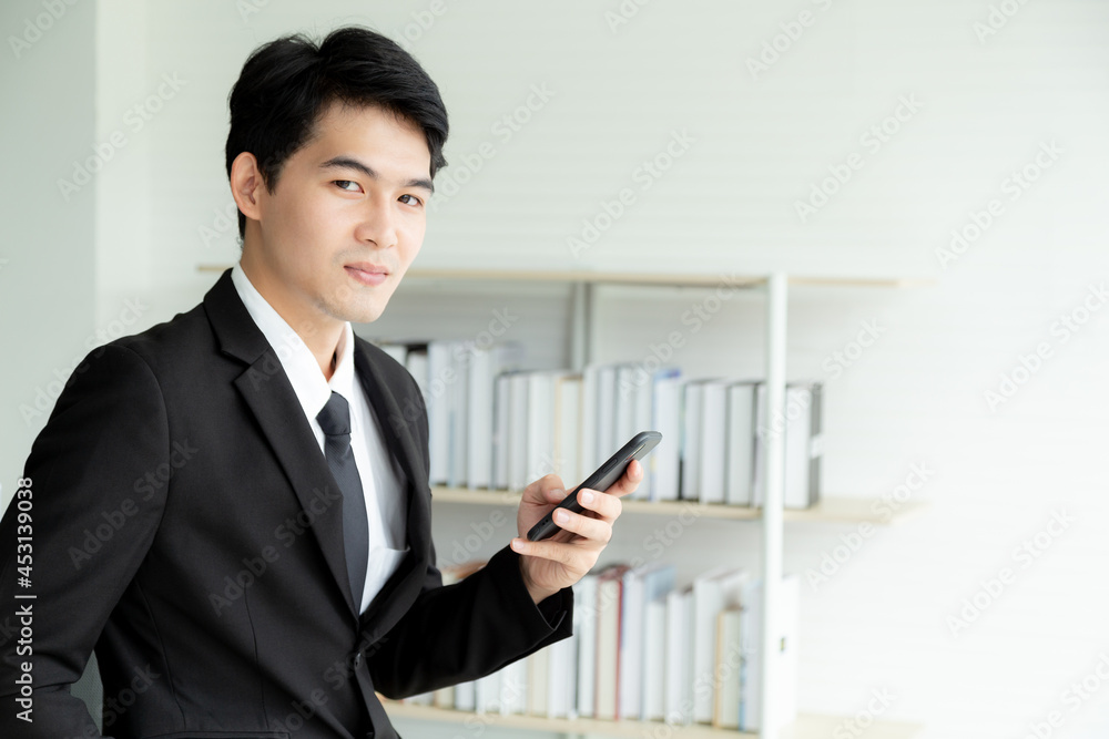 Portrait young businessman in black formal suit using mobile phone online meeting in modern office with business books shelf in the background. Young Asian man holding smartphone for business work. Portrait young businessman in black formal suit using mobile phone online meeting in modern office with business books shelf in the background. Young Asian man holding smartphone for business work.