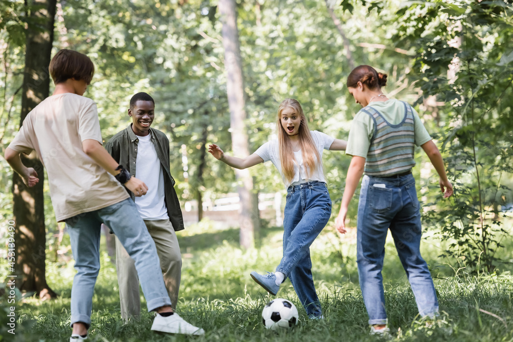 Fototapeta premium Excited girl playing football with multiethnic teenagers in park