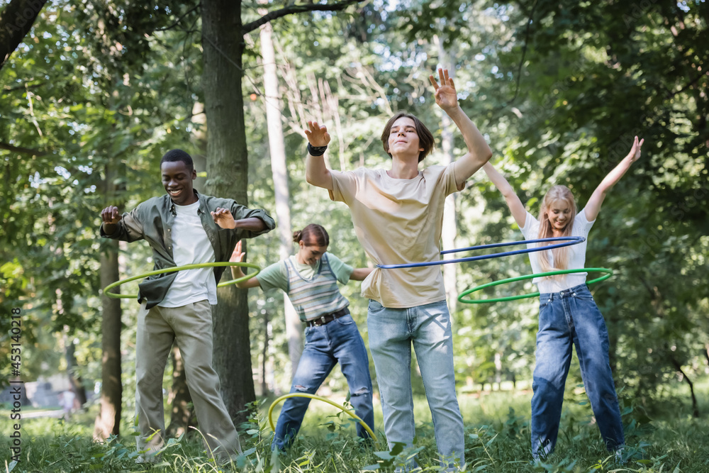 Fototapeta premium Smiling multiethnic teenagers twisting hula hoops in park