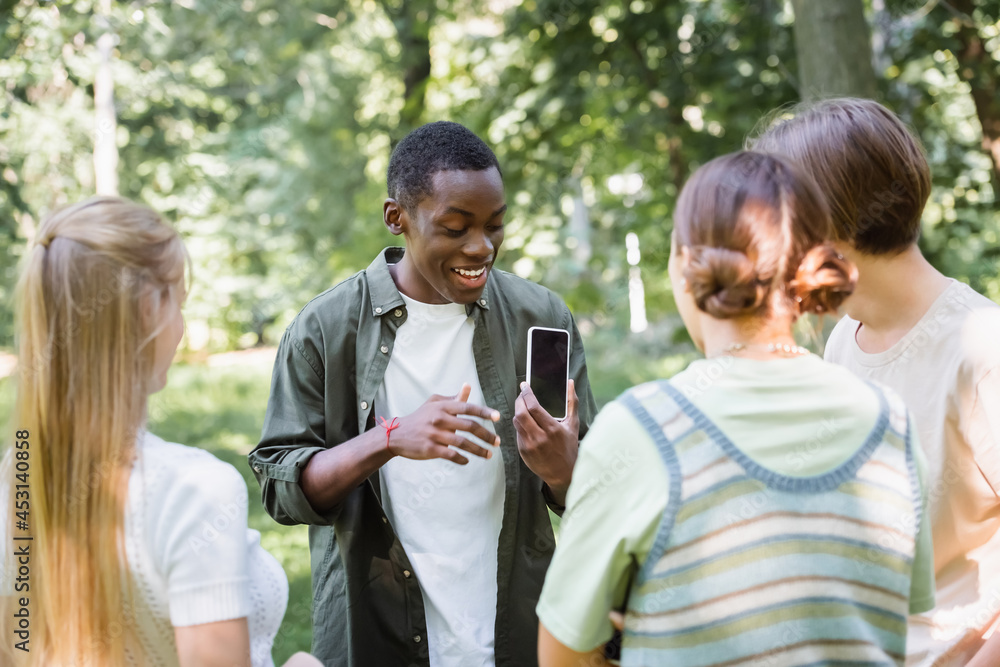 Smiling african american teenager showing smartphone at blurred friends in park