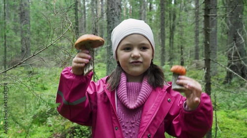 Happy adorable little girl smiles at the camera and shows mushrooms playing eith them in autumn dense forest. Concept of family vacation, quality time together during autumn