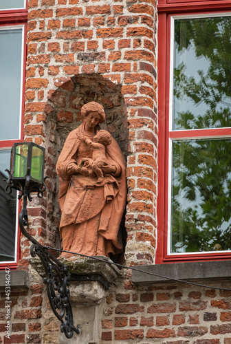 Brugge, Flanders, Belgium - August 4, 2021: Red stone Madonna, mother with child statue in niche between windows of house. Green lantern in front.