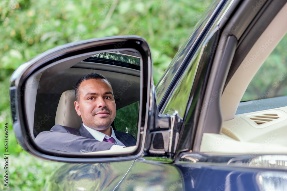 Young man of Middle Eastern appearance in a business suit sitting behind the wheel of a car looks at his reflection in the side rearview mirror. Businessman driving his car. Portrait of a driver