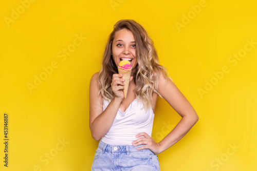 A young excited smiling charming blonde woman with wavy hair in a white top and denim shorts eats fruit ice-cream sorbet in a waffle cone isolated on a bright color yellow background. Summer concept