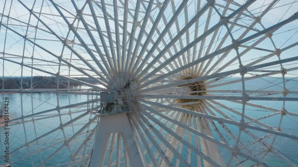 Aerial close-up of a large white Ferris wheel against the background of a blue lake. The mechanism of a large wheel on the background of the horizon line with large mountains-glaciers and snow-capped Aerial close-up of a large white Ferris wheel against the background of a blue lake. The mechanism of a large wheel on the background of the horizon line with large mountains-glaciers and snow-capped