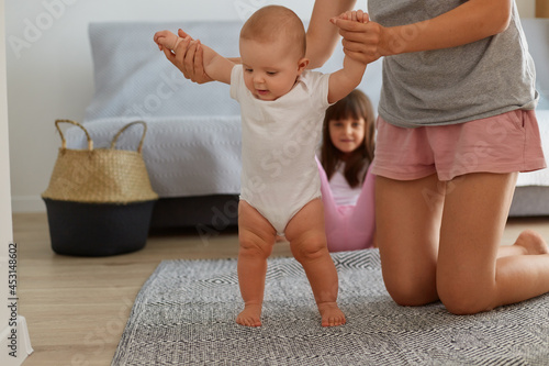 Portrait of beautiful little kid wearing white bodysuit, learn walk with mother, making first steps, family having fun together at home, motherhood and childhood.