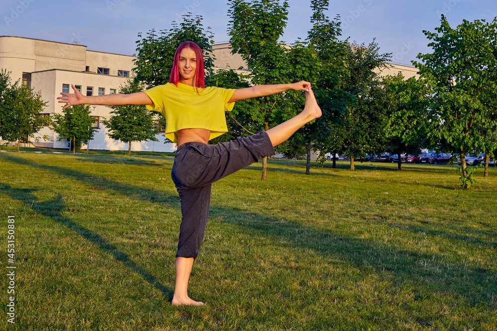 young woman with pink hair trains yoga in a city park in a tree pose, vrishasana