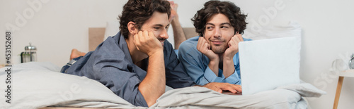 pleased gay couple watching movie on laptop in bedroom, banner