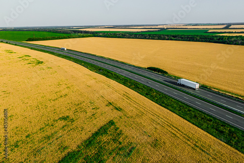 white truck driving on asphalt road aalong the yellow fields of wheat at sunset. seen from the air. Aerial view landscape. drone photography. cargo delivery © drotik