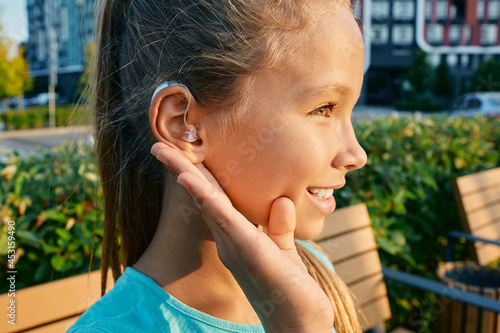 Smiling female child with a hearing aid behind the ear holds hand near her ear for listening environment outdoor, having full life photo