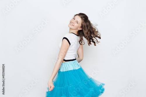 Cheerful teenager girl with curly hair wearing blue fluffy skirt and white t shirt posing on white background. Emotions, happiness, motion