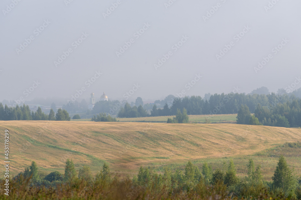 Hilly Belarusian landscape. Foggy morning. Sunrise over a field with trees and a church on the horizon.