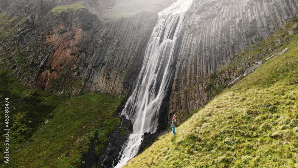 Happy female traveler admires a giant mountain waterfall with her hands raised up standing on a picturesque green slope, aerial view Happy female traveler admires a giant mountain waterfall with her hands raised up standing on a picturesque green slope, aerial view