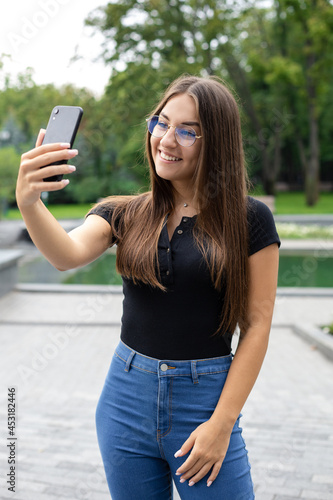 Online education, technology and Internet concept — A smiling European dark-haired girl in casual clothes stands in a park in the summer talking to her classmates by video link in class.
