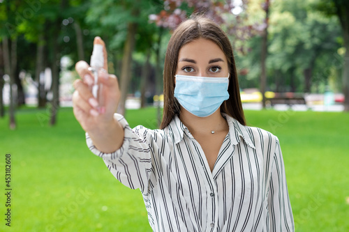 COVID-19, pandemic, protective equipment, hand treatment — A woman wearing a protective mask with dark hair in a light-colored shirt directed antiseptic into a camera in a park in sunny summer weather