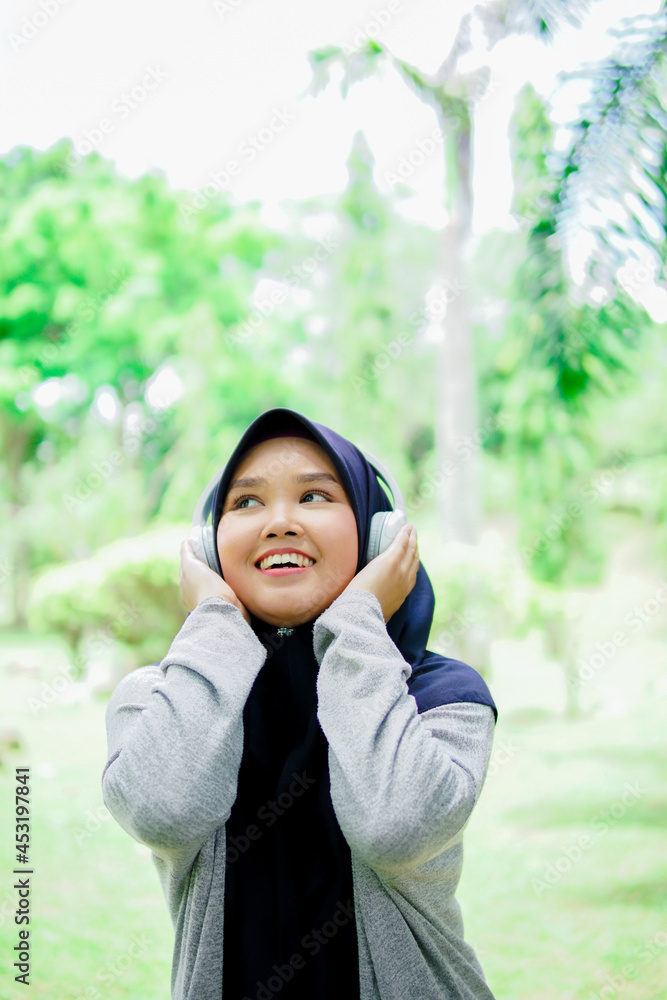 Portrait of an attractive and beautiful young Asian Muslim woman listening to music with headphones in a park Portrait of an attractive and beautiful young Asian Muslim woman listening to music with headphones in a park