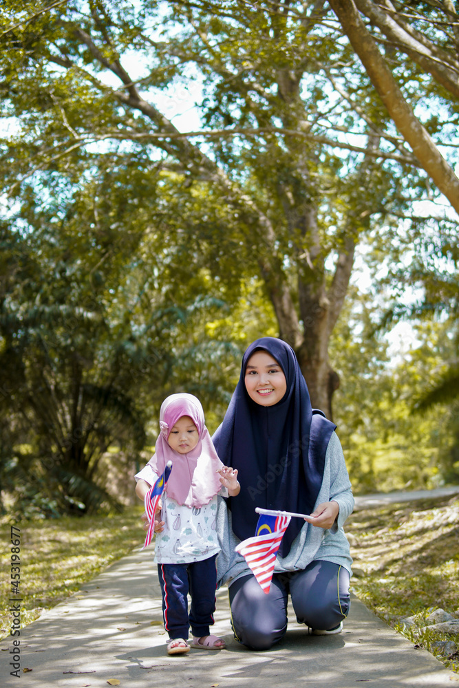 Fototapeta premium Beautiful young mother with her daughter celebrating Malaysia independence day by raising flag