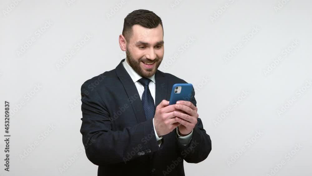 Smiling happy young adult bearded businessman in black official style suit standing holding smart phone, browsing internet, reading good news. Indoor studio shot isolated on gray background. Smiling happy young adult bearded businessman in black official style suit standing holding smart phone, browsing internet, reading good news. Indoor studio shot isolated on gray background.