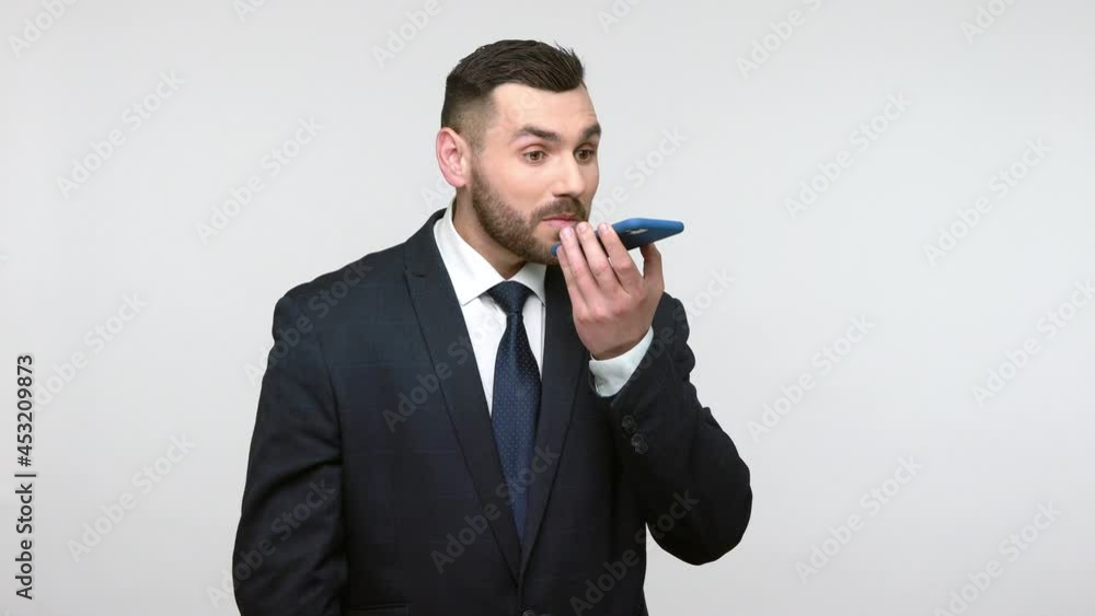Virtual assistant. Young adult bearded businessman in black suit talking to cellphone giving voice command to digital ai technology application. Indoor studio shot isolated on gray background. Virtual assistant. Young adult bearded businessman in black suit talking to cellphone giving voice command to digital ai technology application. Indoor studio shot isolated on gray background.