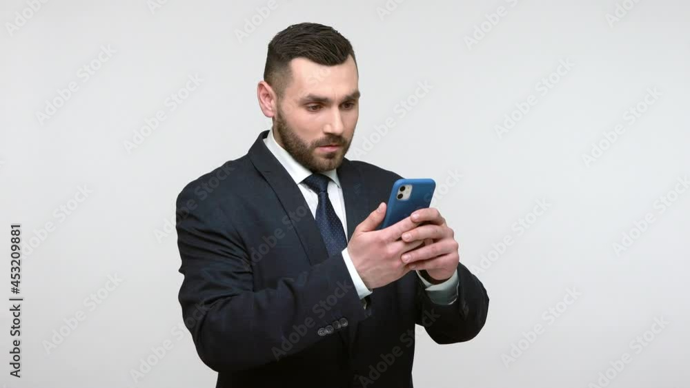 Amazed bearded businessman in black official style suit satnding with smart phone in hands, being shocked of surprising content, saying wow. Indoor studio shot isolated on gray background. Amazed bearded businessman in black official style suit satnding with smart phone in hands, being shocked of surprising content, saying wow. Indoor studio shot isolated on gray background.