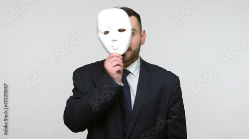 Confident handsome bearded businessman in black official style suit putting off mask showing different emotions, duplicity, hypocrisy. Indoor studio shot isolated on gray background.