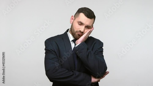 Lazy bored sleepy man in suit standing leaning on hand, looking at camera with bored indifferent expression, exhausted of tedious communication. Indoor studio shot isolated on gray background.