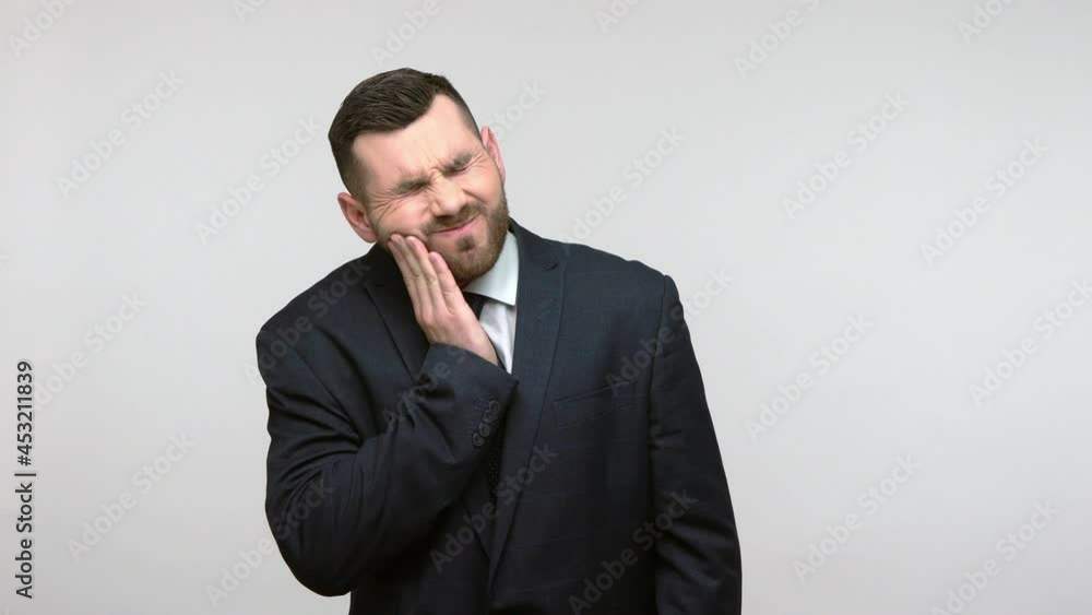 Dental problems. Unhealthy bearded businessman in official style suit touching sore cheek, suffering terrible toothache, horrible pain from cavities. Indoor studio shot isolated on gray background.