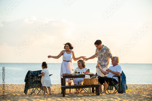 Happy Asian family on holiday vacation. Group of multi generation family little girl with parents and grandparents relax and enjoy with dinner party and dancing together on the beach at summer sunset.