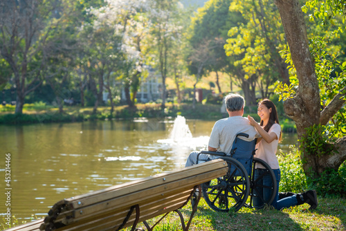 Happy Asian woman caring and hugging senior man grandfather sitting on wheelchair in the park. Elderly retired male relax and enjoy outdoor leisure activity with daughter. Family relationship concept.