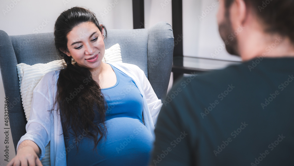 Beautiful young pregnant woman sitting on bed with loving and caring husband trying to connect with baby by touching and feeling stomach at home Beautiful young pregnant woman sitting on bed with loving and caring husband trying to connect with baby by touching and feeling stomach at home