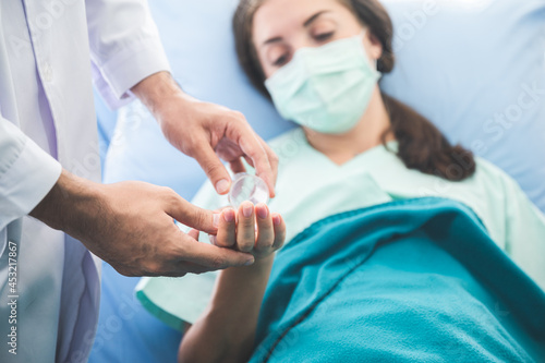 Young woman suffering with corona virus infection lying on hospital bed with mask sleeping and resting while doctor taking notes of progress and writing on clipboard in background wearing ppe kit