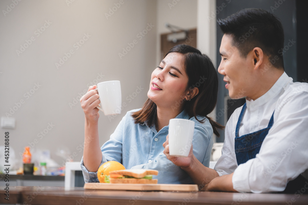 Cheerful and romantic young husband wearing apron drinking coffee with beautiful wife with sandwich and fruits at kitchen table while looking at each other Cheerful and romantic young husband wearing apron drinking coffee with beautiful wife with sandwich and fruits at kitchen table while looking at each other