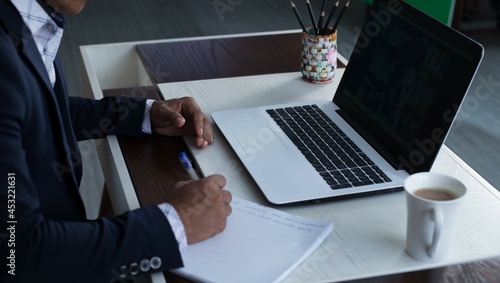 Young adult male writing down notes on a notebook. Unrecognizable businessman with laptop computer on the desk  working. 