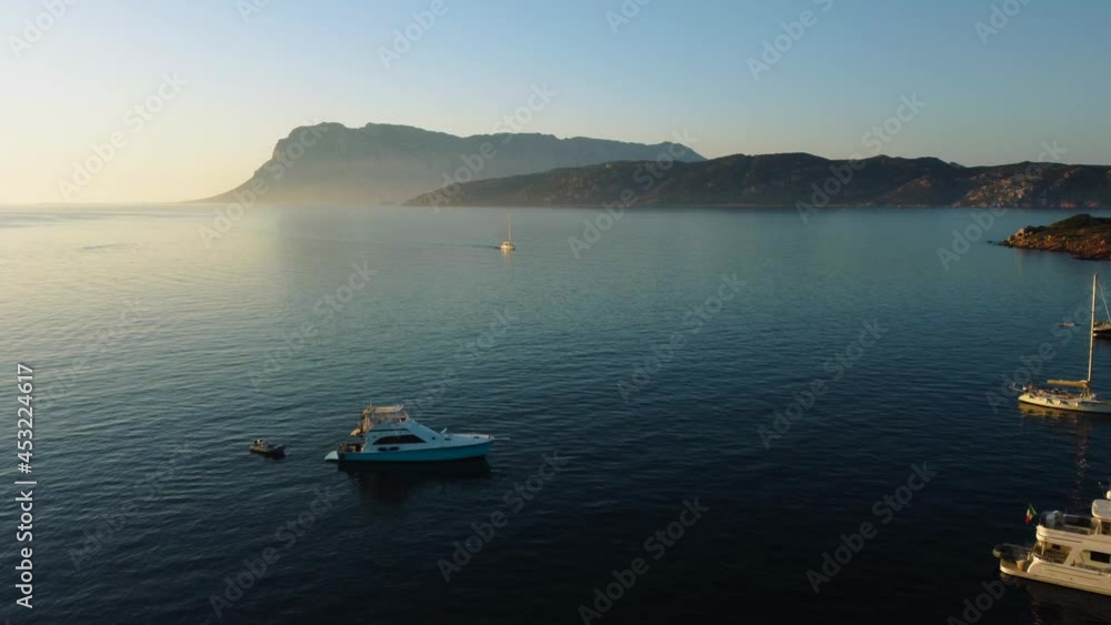 Flying over sailing boats on the tourist vacation island Sardinia in Italy at sunset at an idyllic sandy beach bay with clear blue turquoise and calm water close to Olbia. Aerial drone air view flight Flying over sailing boats on the tourist vacation island Sardinia in Italy at sunset at an idyllic sandy beach bay with clear blue turquoise and calm water close to Olbia. Aerial drone air view flight