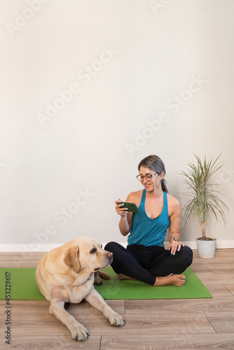 A girl in a blue T-shirt is resting after a home workout with a phone in her hands, her dog is lying nearby