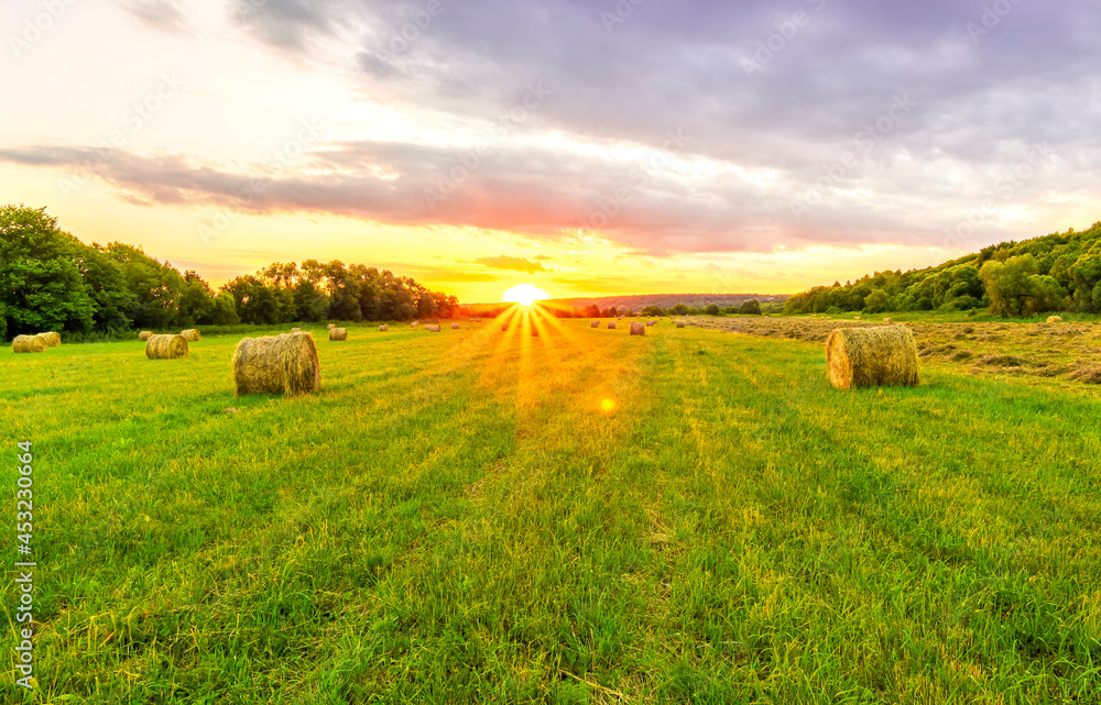 Obraz premium Scenic view at beautiful sunset in green shiny field with hay stacks, bright cloudy sky, country road and golden sun rays with glow, summer valley landscape