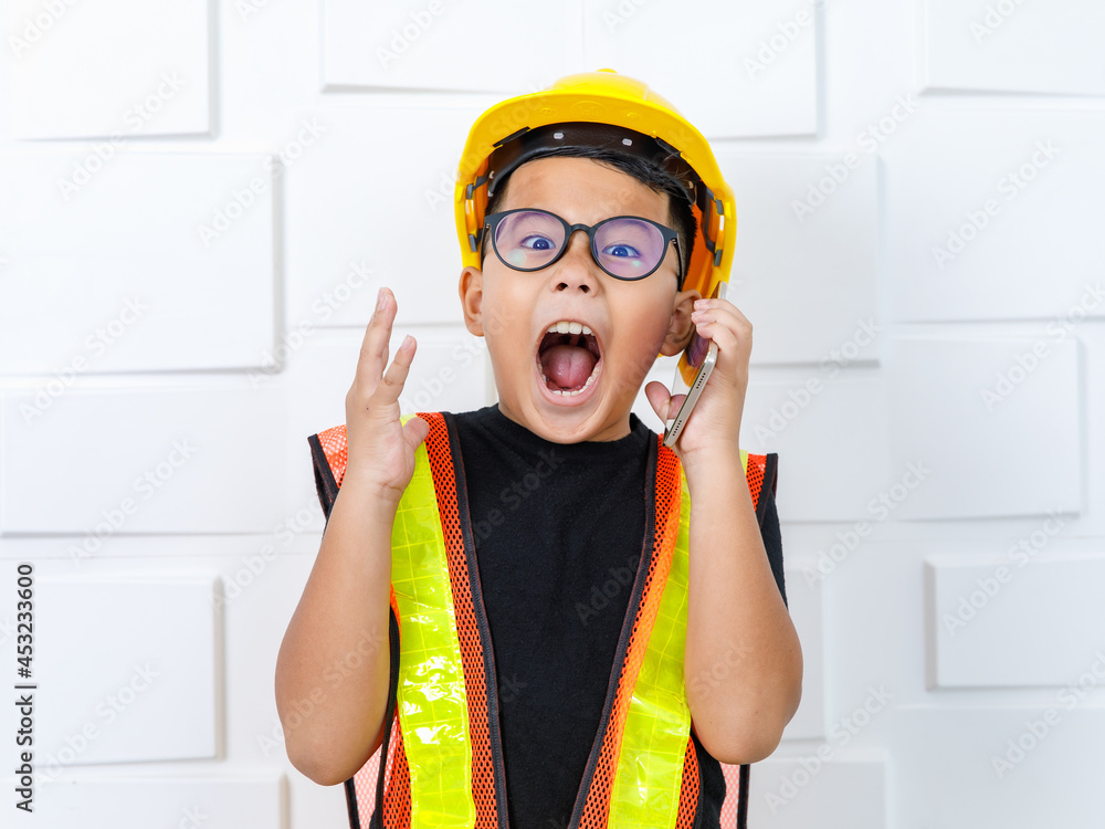 Young Asian boy wearing glasses, black shirt, yellow safety vest and helmet standing near white wall and shout out loud as shocked or exciting on unexpected contacting info from smartphone calling Young Asian boy wearing glasses, black shirt, yellow safety vest and helmet standing near white wall and shout out loud as shocked or exciting on unexpected contacting info from smartphone calling