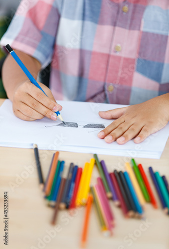 Asian kid on pink checked shirt sitting on class table full of color pencils to study art course and carefully draw and shade picture of black glasses on white paper by blue pencil