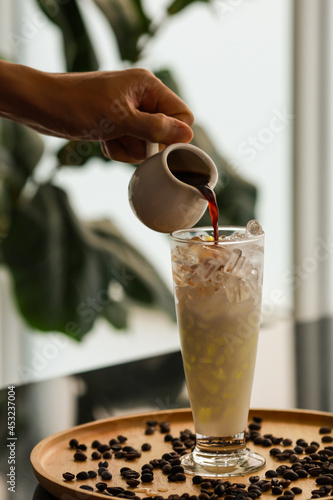Black coffee in small white jug was holded and poured by barista left hand into tall glass with cold ice and milk placed on brown wooden tray full of coffee beans with blurred background