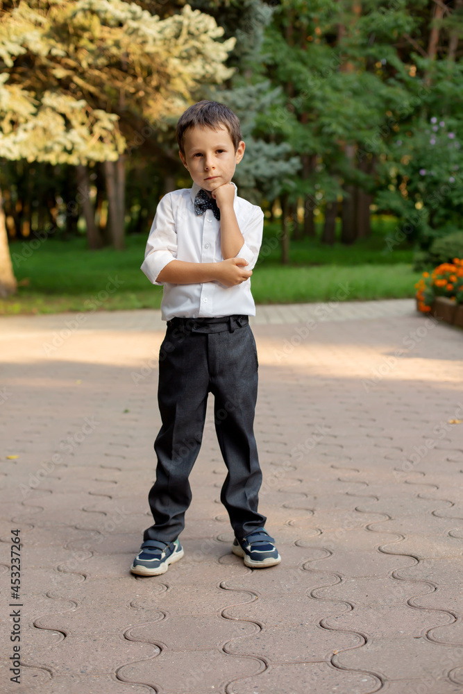 a beautiful little boy in a white shirt and gray trousers a schoolboy with a bow around his neck is standing in the school yard a beautiful little boy in a white shirt and gray trousers a schoolboy with a bow around his neck is standing in the school yard
