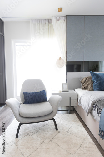 White armchair and carpet next to cozy king size bed with white bedding in fashionable bedroom interior. © bennnn