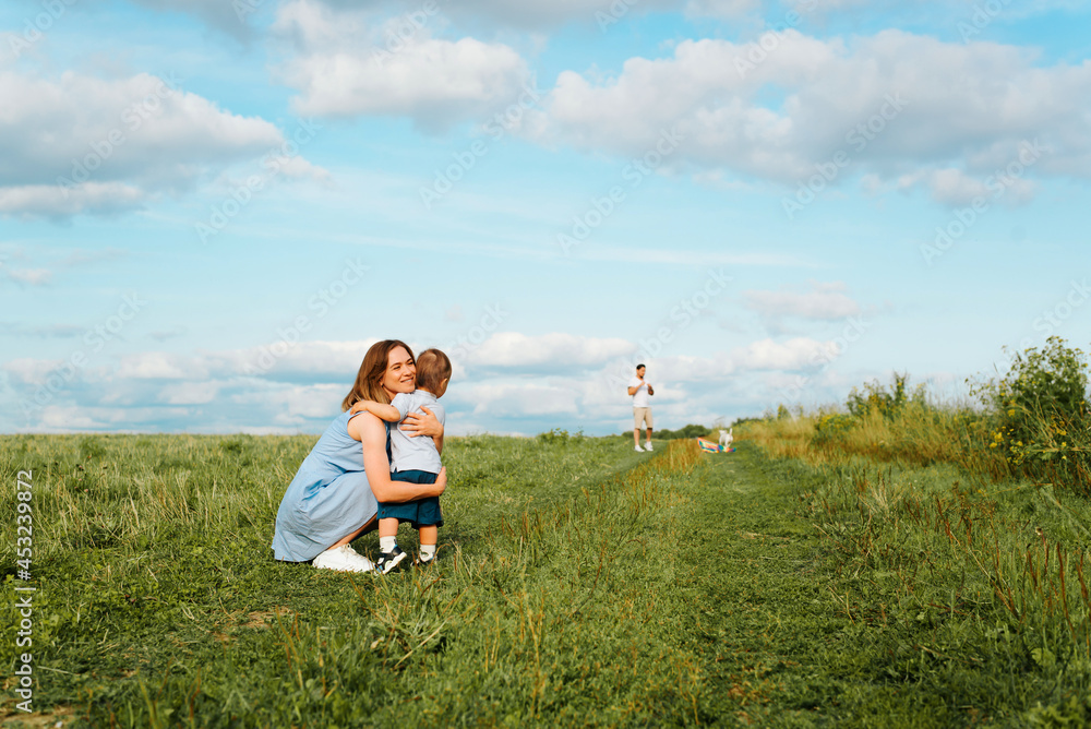 Family outdoor recreation. Smiling young mother hugs small child in field, dad with dog is standing in distance out of focus. Motherly love, spending time together in nature Family outdoor recreation. Smiling young mother hugs small child in field, dad with dog is standing in distance out of focus. Motherly love, spending time together in nature