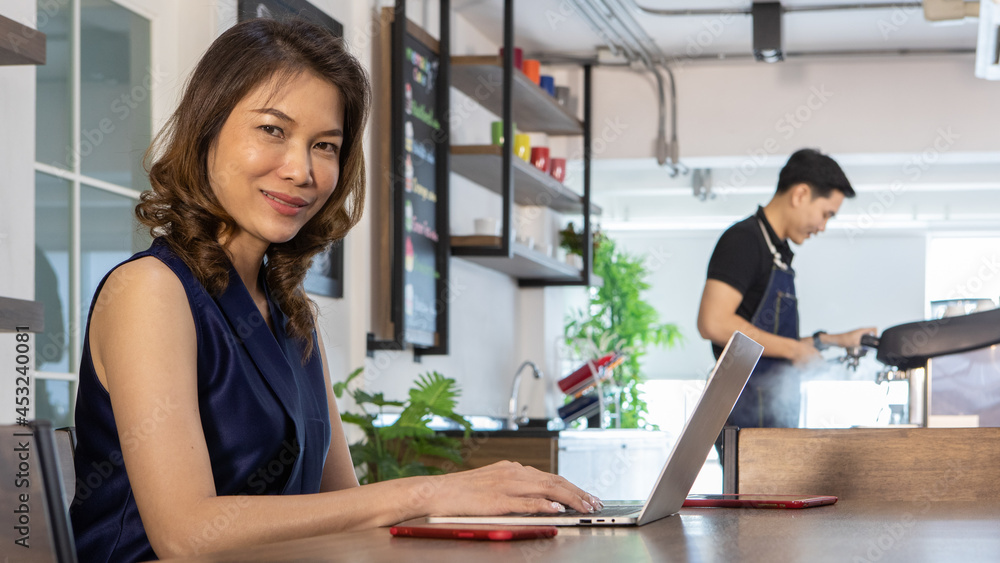 Pretty young Asian woman freelancer in dark tank top sitting near counter of barista in cafe on afternoon, turn to smile while typing on laptop as happy to work online business Pretty young Asian woman freelancer in dark tank top sitting near counter of barista in cafe on afternoon, turn to smile while typing on laptop as happy to work online business