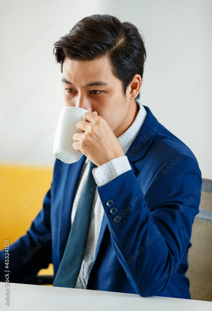 Asian youn businesman in blue suit sitting in office and holding coffee cup with easy and relax pose Asian youn businesman in blue suit sitting in office and holding coffee cup with easy and relax pose
