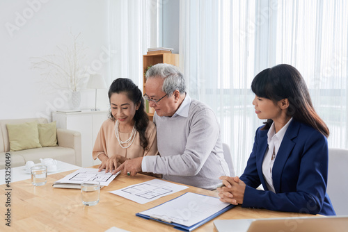Smiling senior man and woman meeting with real estate broker and looking and building plans when choosing new house