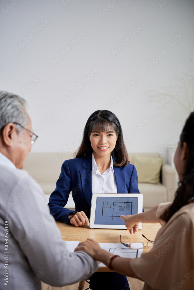 Pretty smiling young real estate agent showing tablet computer with building plan to senior couple who want to buy new house Pretty smiling young real estate agent showing tablet computer with building plan to senior couple who want to buy new house