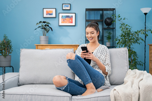 Smiling beautiful teenage girl is sitting on sofa in living room holding phone, girl is sending messages to friends, writing a blog post, replying to emails, chatting, tapping on smartphone screen