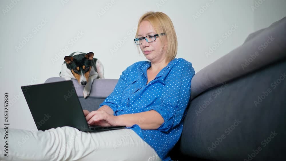 An adult blond woman in glasses working on a laptop from home while sitting on the floor with her cute Basenji dog. An adult blond woman in glasses working on a laptop from home while sitting on the floor with her cute Basenji dog.