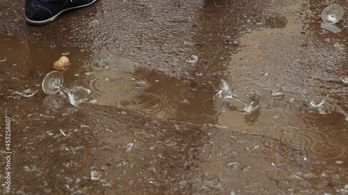 Broken glasses and cork lying in puddle on the road. It raining. Military man in shoes, pants and raincoat pass by. Oficer pushes broken dishes with foot. Territory occupation, hostilities in city. photo