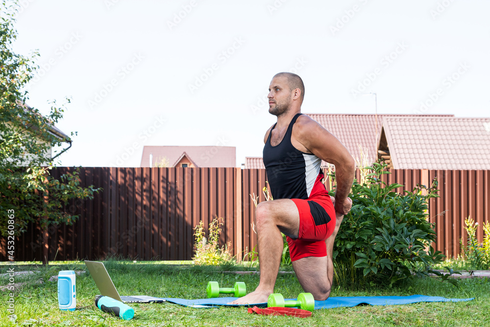 The young man goes in for sports at home in  backyard in summer day. Young sportsman with blond  hair  doing  lunges , stretching on mat, there is a laptop, a buttle  and rubber for sports nearby